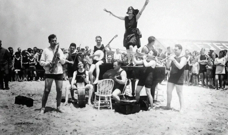 A black-and-white photo of a jazz band performing on a beach, with musicians playing instruments and a woman standing on a piano with arms raised while a crowd watches in the background.