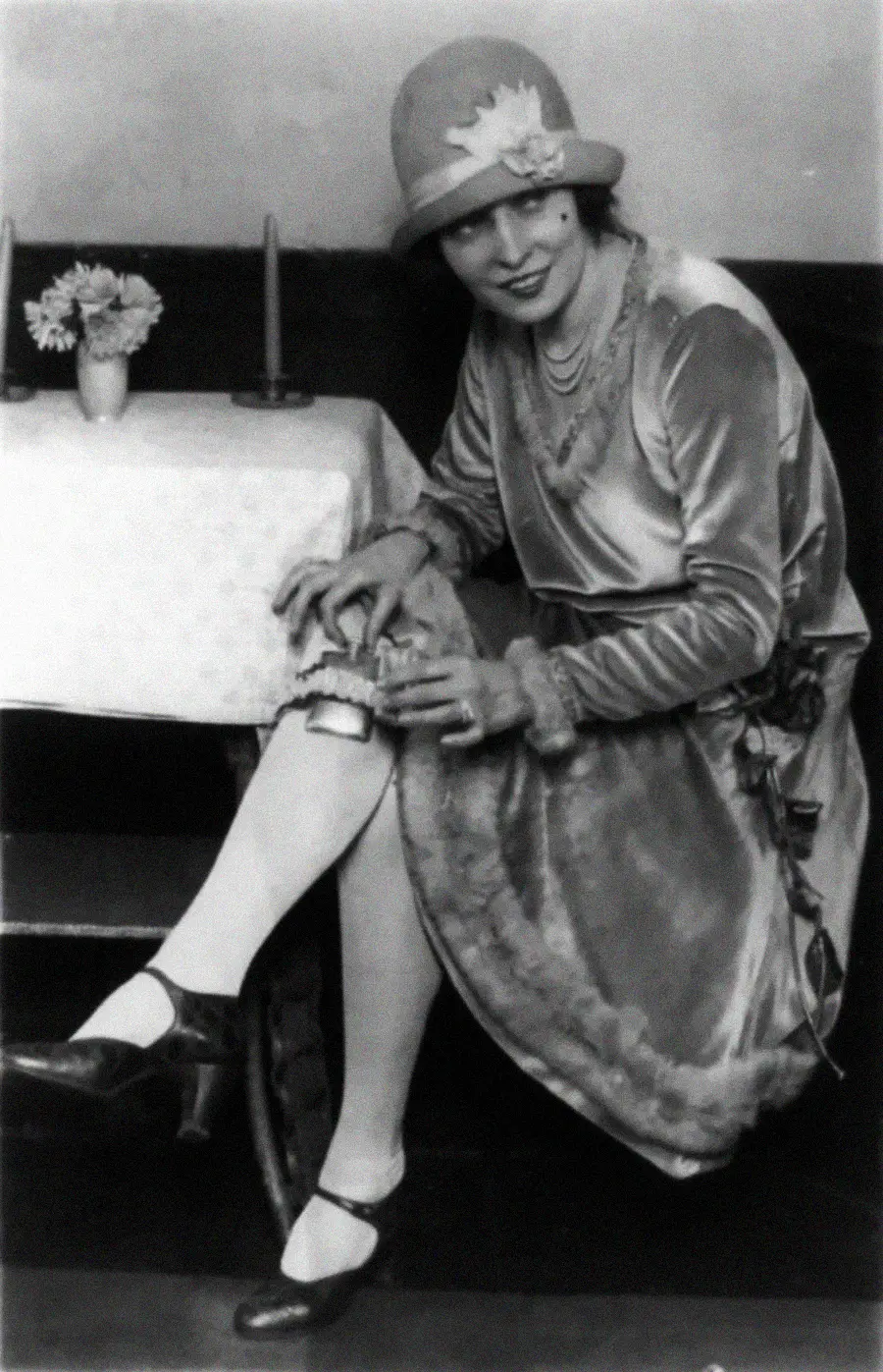 A woman in 1920s attire sits beside a table, lifting her dress to reveal a flask strapped to her thigh. She wears a cloche hat, a velvet dress with fur trim, and heels, and smiles at the camera.
