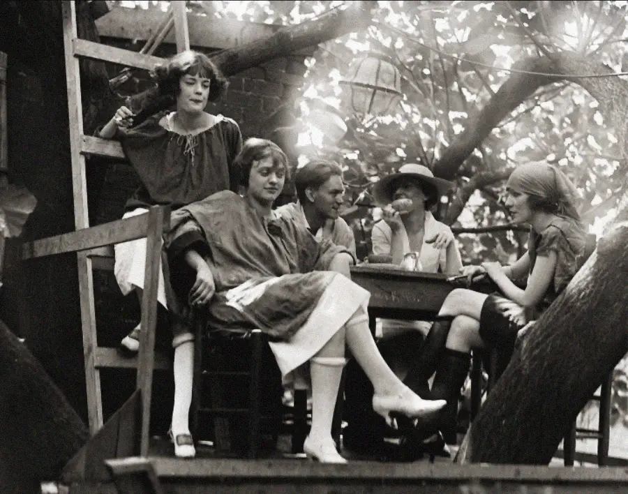Five women in 1920s-style clothing sit and talk around a small table in a treehouse, surrounded by branches and dappled sunlight. Some are relaxed, while others appear engaged in conversation.