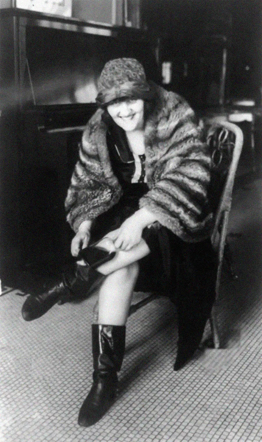 A woman in a fur coat and cloche hat sits on a wicker chair, smiling as she adjusts her shoe. The black-and-white photo has a vintage feel, likely from the early 20th century, with a tiled floor and a piano in the background.