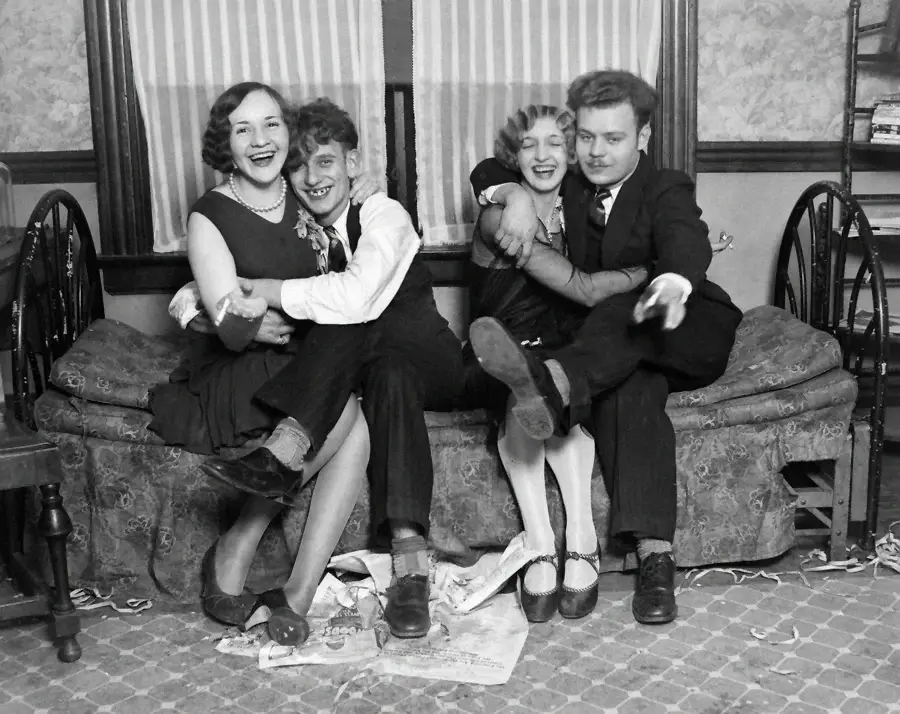 Four young adults sit closely together on a sofa, smiling and laughing. Two women and two men, dressed in vintage clothing, pose playfully with arms around each other in a cozy, old-fashioned living room.