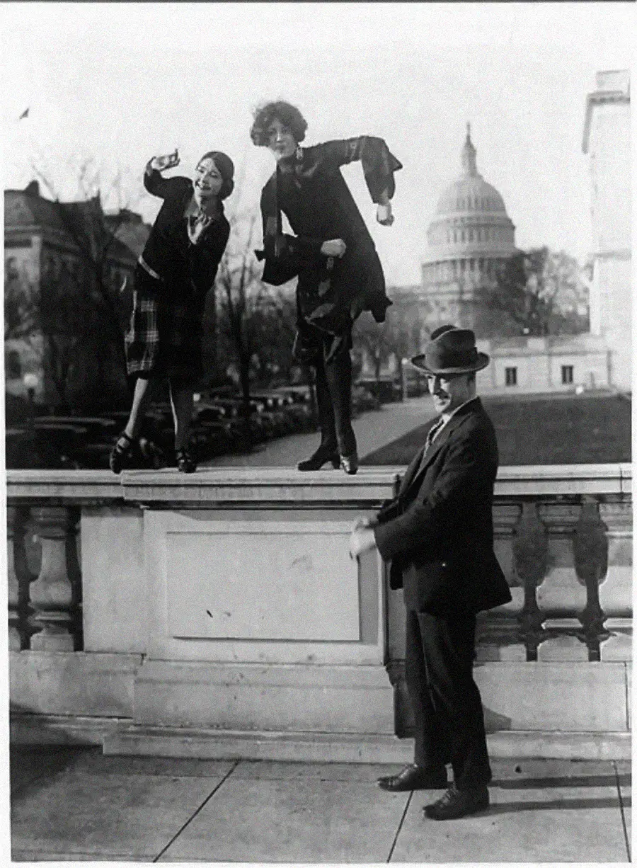 Two women in 1920s clothing pose playfully on a railing, one appearing to take a selfie with a camera, while a man in a hat stands below them. The U.S. Capitol building is visible in the background.