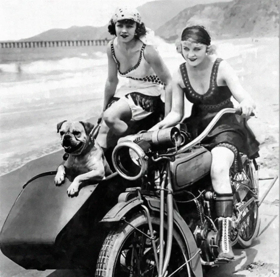 Two women in vintage swimsuits pose on a beach; one sits on a motorcycle with a dog in its sidecar, while the other kneels beside them. The background shows the ocean and distant hills.