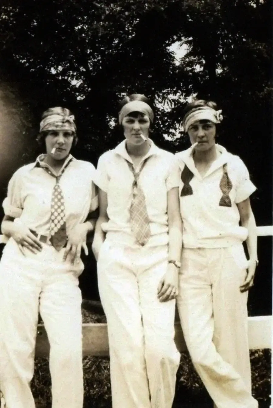 Three women stand side by side outdoors, wearing white shirts, pants, neckties, and headscarves. They pose casually in front of a wooden fence with trees in the background, looking at the camera.