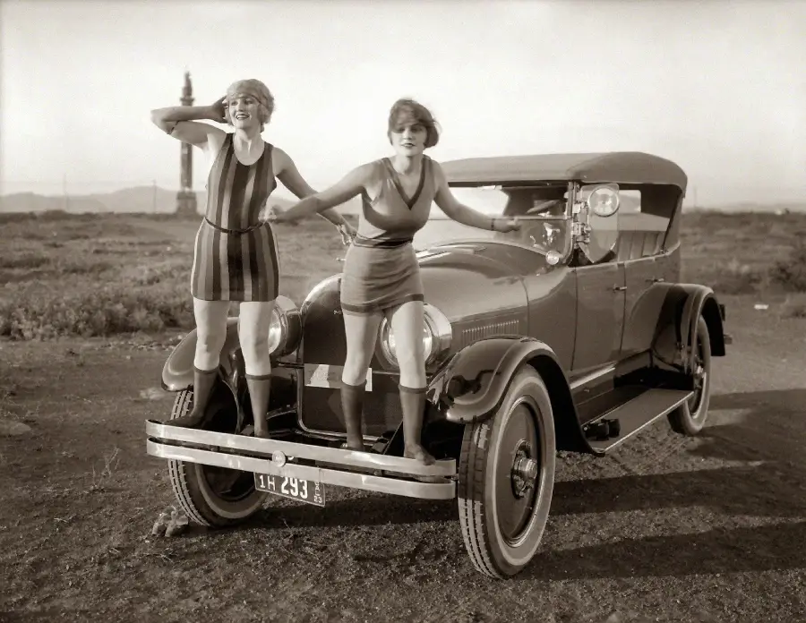 Two young women in 1920s-style dresses stand on the front bumper of a vintage car in an open field, smiling and striking playful poses. A tall structure is visible in the distant background.