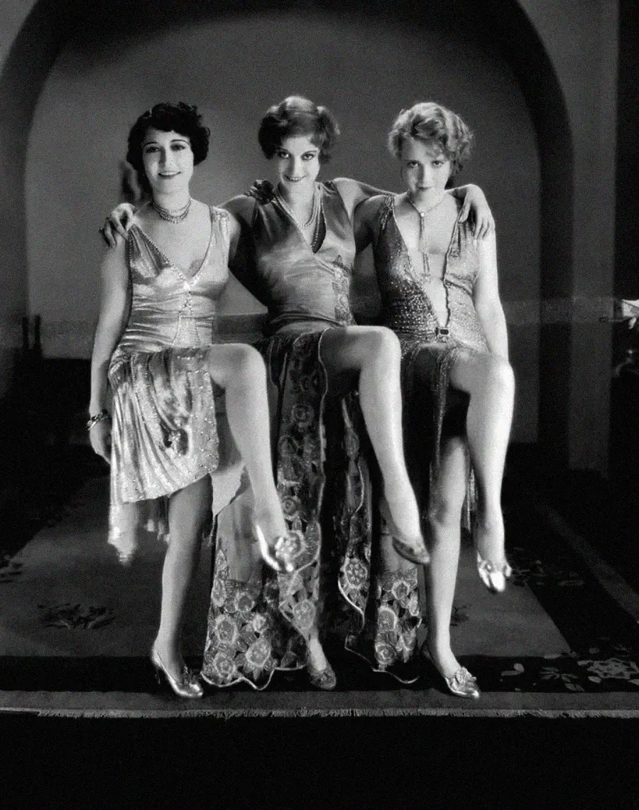 Three women in 1920s flapper dresses sit on a ledge indoors, smiling and raising one leg each in a playful pose. The image is black and white and has a vintage, glamorous atmosphere.