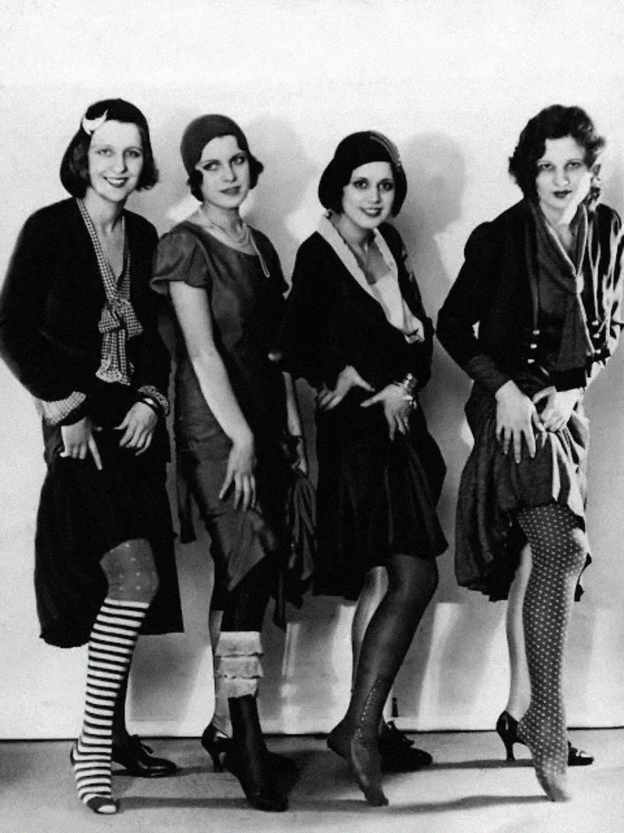Four women in 1920s-style dresses and various patterned stockings pose with one leg forward, smiling and looking at the camera against a plain backdrop.
