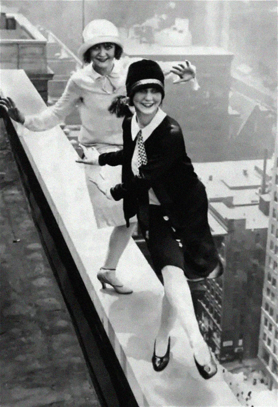 Two women in 1920s clothing balance and smile on the edge of a tall building rooftop, with a cityscape visible far below them.