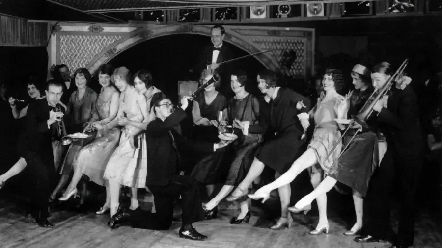 A group of people in 1920s attire dance and kick their legs in a lively line, while musicians play instruments and a man in a tuxedo stands in the background, evoking a festive, Jazz Age party scene.