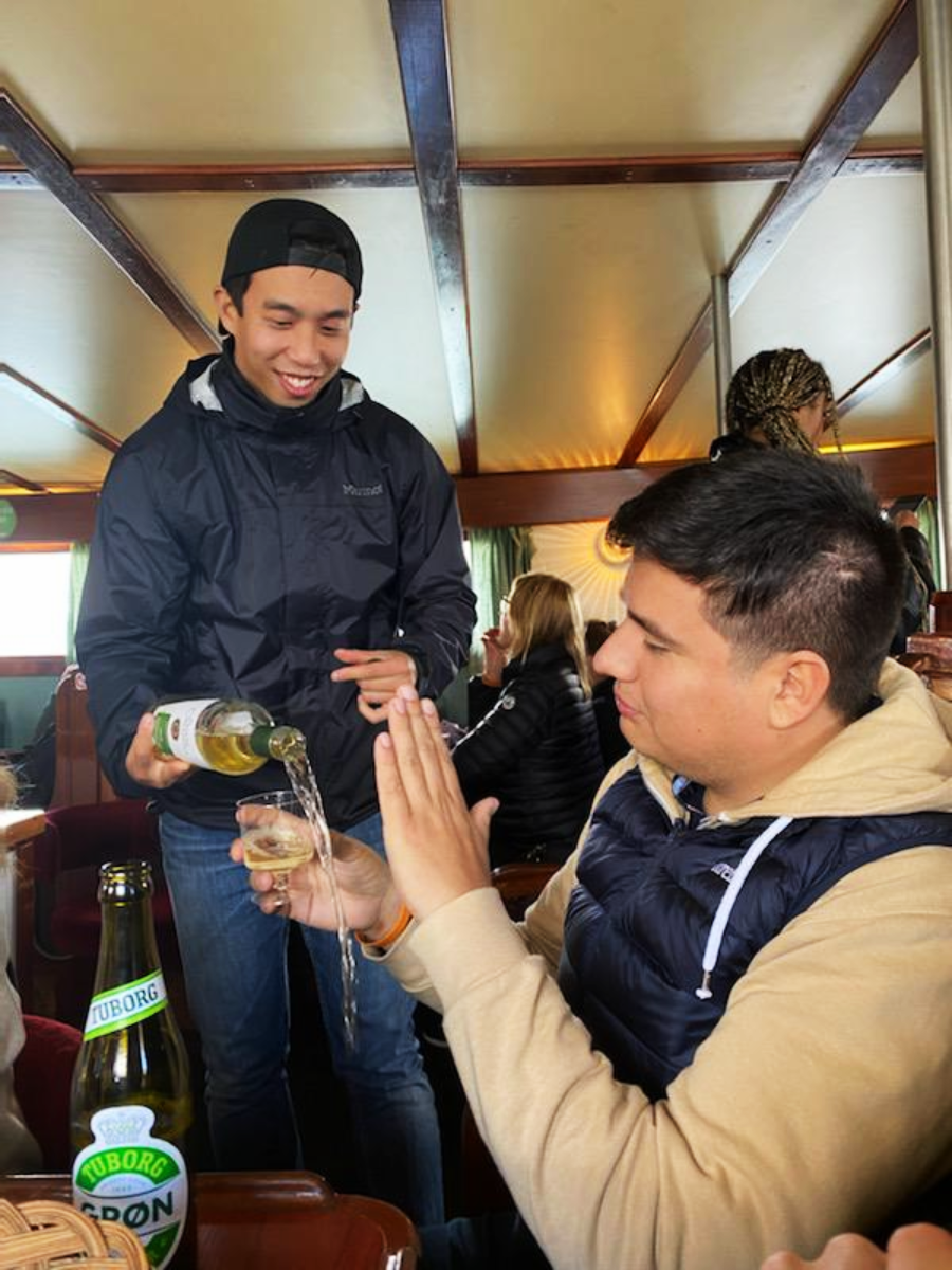 A young man in a black jacket laughs as he accidentally overfills a glass with beer, spilling it onto another man's hand. They are sitting in a cozy, wooden interior with other people in the background.