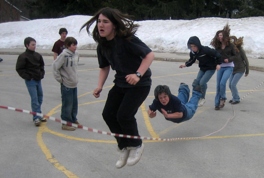 A group of kids play jump rope on an outdoor court with snow in the background; one child is airborne horizontally as others jump or watch.