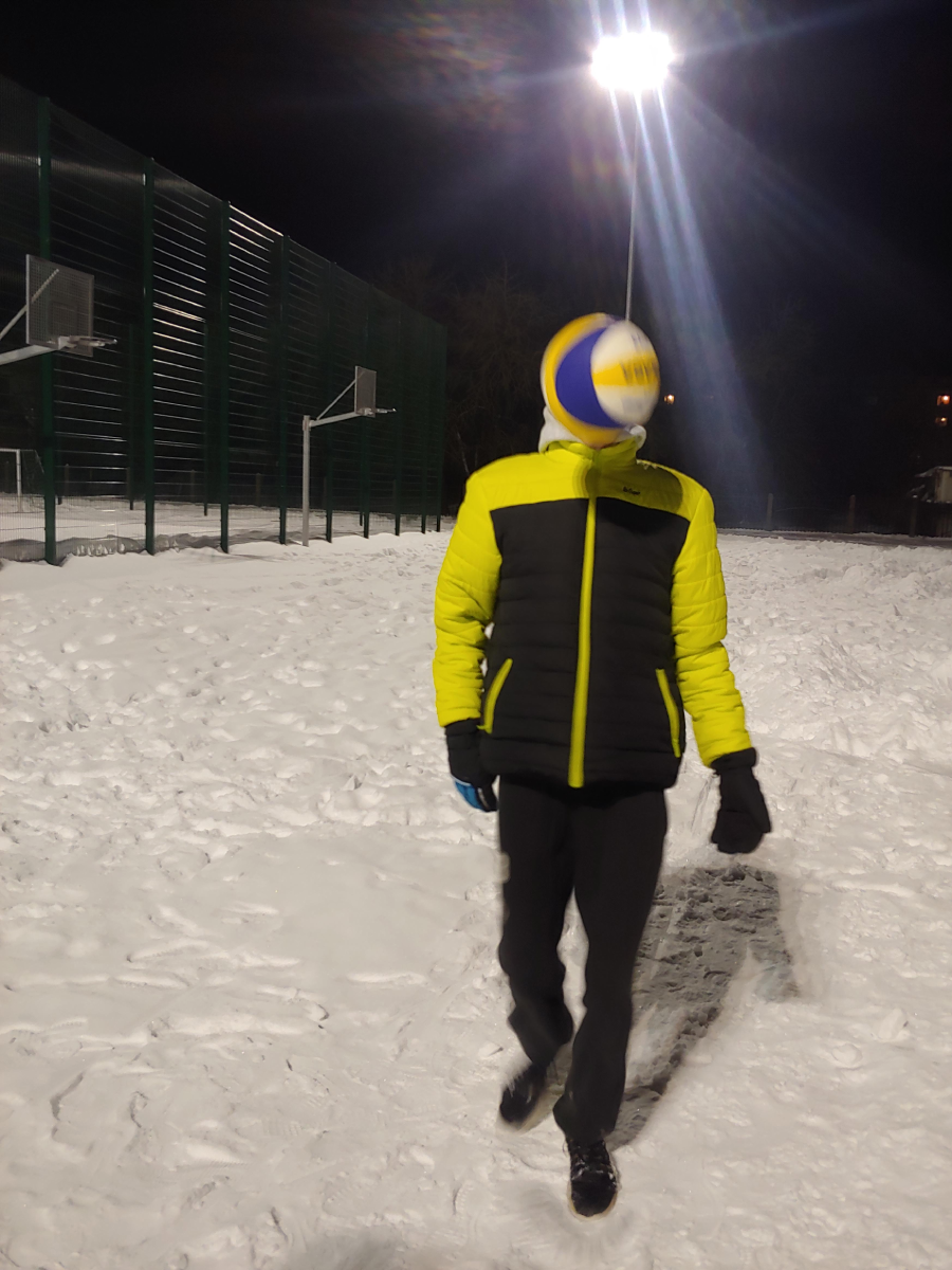 A person in a black and yellow jacket walks on a snowy outdoor court at night, with a volleyball caught mid-air, obscuring their face. Bright floodlights illuminate the scene.