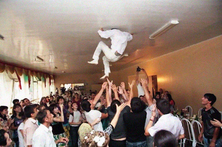 A group of people at a party cheer and raise their hands as one person, dressed in white, is thrown into the air and touches the ceiling with their hands and feet.