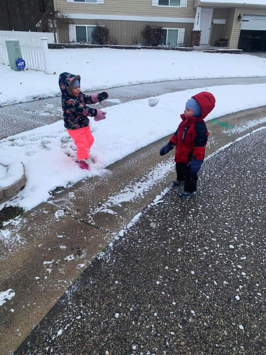 Two young children play in a snowy suburban neighborhood. One child, wearing bright pink pants and a floral jacket, throws a snowball towards the other child, who is dressed in a red snowsuit and winter hat.