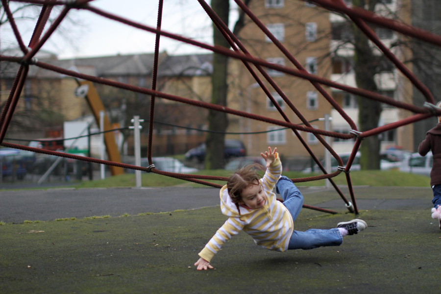 A young child in a striped hoodie is falling onto the ground from a red rope climbing structure at a playground, with one hand out to catch themselves. Buildings and trees are visible in the background.