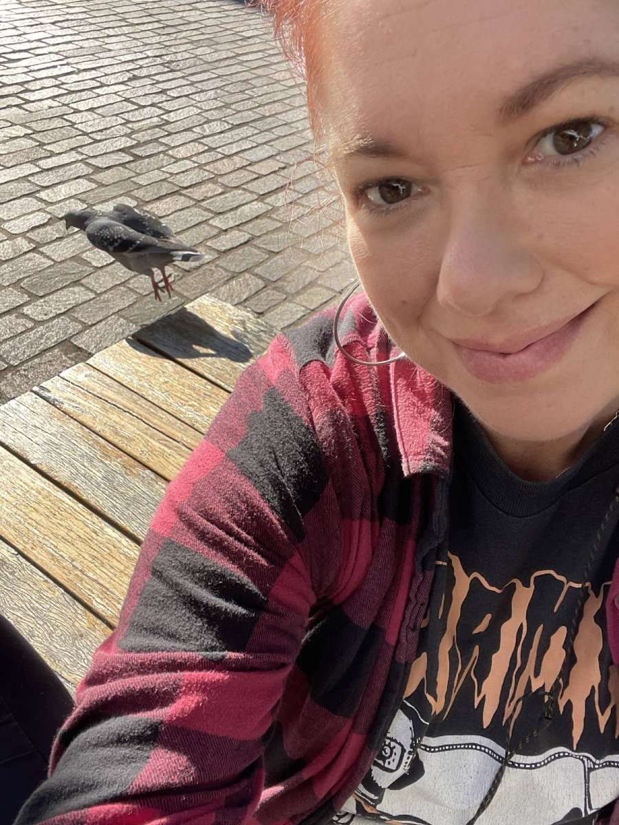 A person with reddish hair, wearing a plaid shirt and graphic tee, smiles for a selfie on a sunny day while sitting on a wooden bench; a pigeon walks on the pavement nearby.
