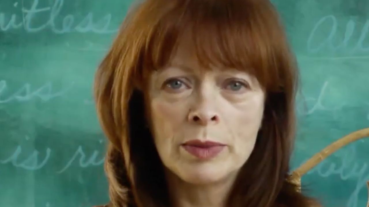 A woman with straight red hair and bangs stands in front of a green chalkboard with cursive writing on it, looking directly at the camera with a neutral expression.