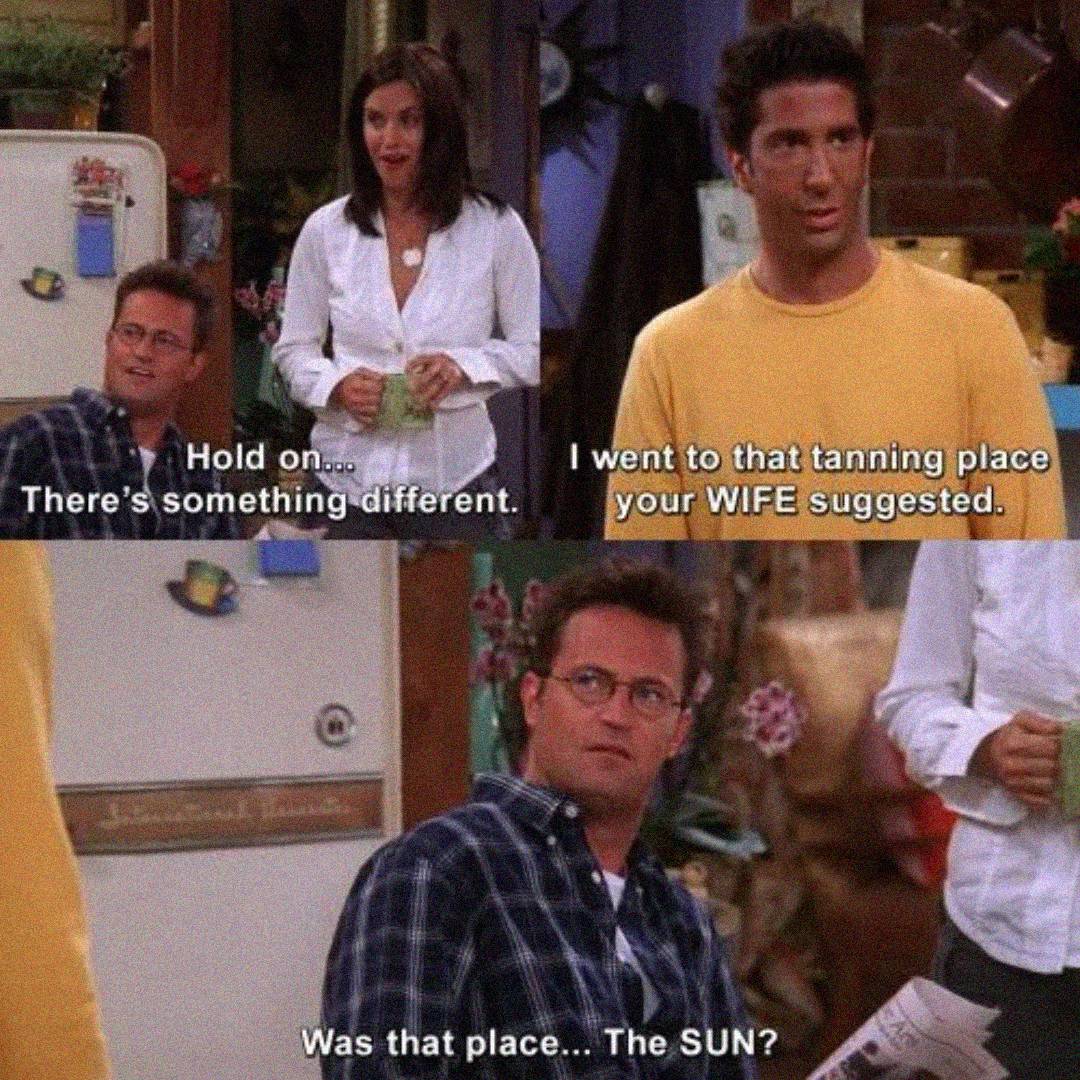 Three people stand in a kitchen. One man in a yellow shirt says he went to a tanning place suggested by another man's wife. The second man sarcastically asks, "Was that place... the SUN?