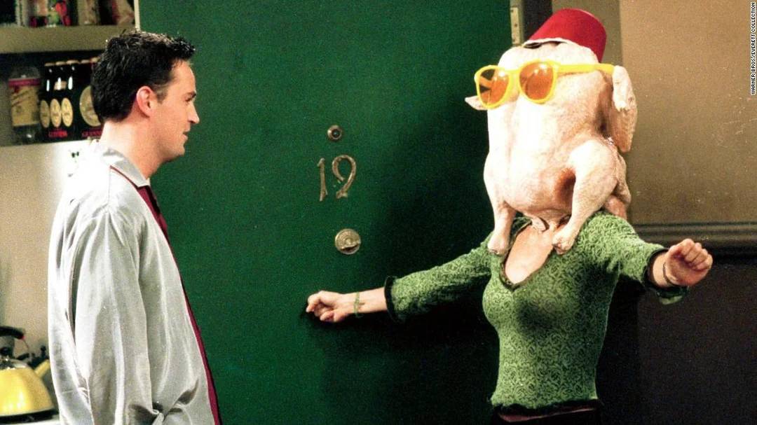 A man stands in front of a green door marked "19" while a woman, wearing a cooked turkey with sunglasses and a red hat on her head, spreads her arms wide in a comedic pose.