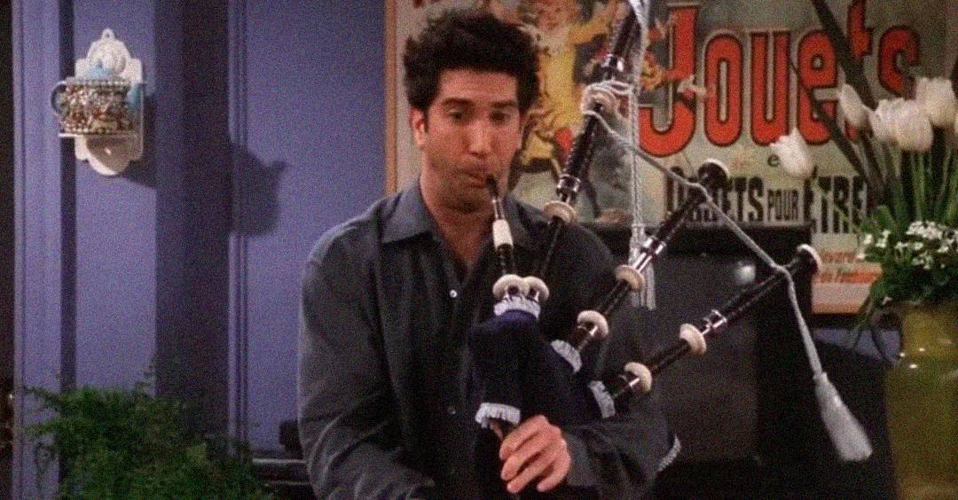 A man with dark hair, wearing a dark shirt, plays the bagpipes indoors. Behind him, there is a vintage poster, some plants, and flowers decorating the room.