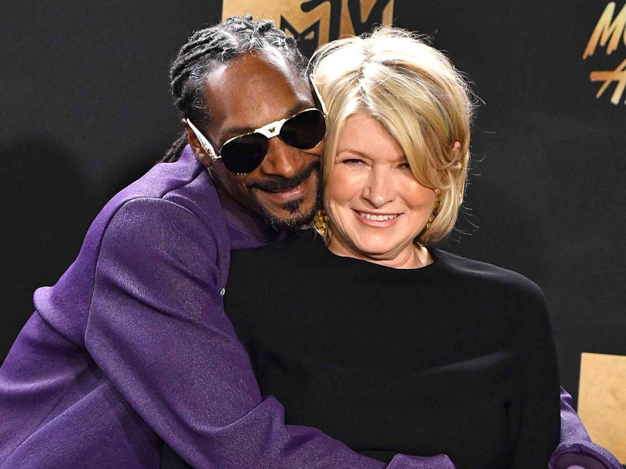 A man in a purple suit and sunglasses hugs a smiling woman in a black top from behind. Both are posing together at an event with a dark background and gold text.