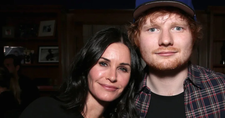 A woman with long dark hair and a man with reddish hair and a beard stand close together, smiling slightly at the camera in a warmly lit indoor setting.