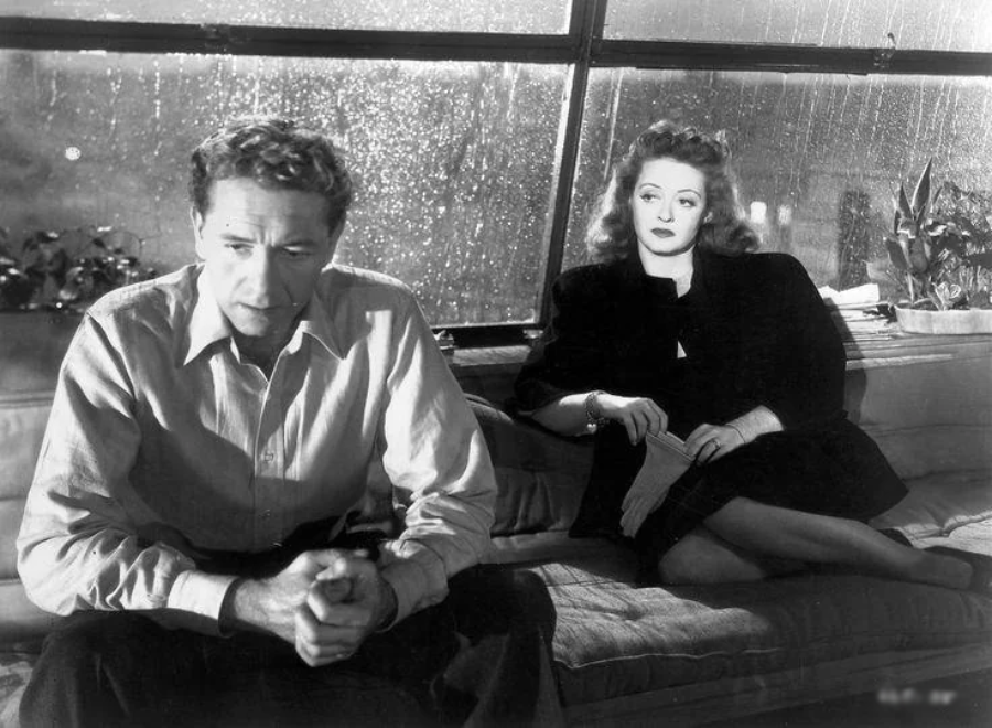 A man sits on a couch looking thoughtful and tense, while a woman in a dark dress sits beside him, watching him intently. Rain streams down the large window behind them, suggesting a somber mood.