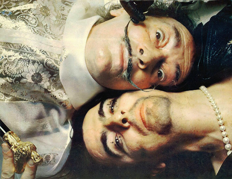 Two people lie close together, heads touching. One wears a lace outfit, sports an upturned mustache, and holds a cane; the other wears a pearl necklace and has dramatic makeup with sparkling accents near the eyes.