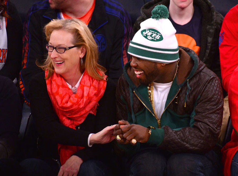 A woman with blonde hair and glasses laughs with a man wearing a green and white New York Jets beanie and brown jacket as they sit together in a crowd at a sports event.
