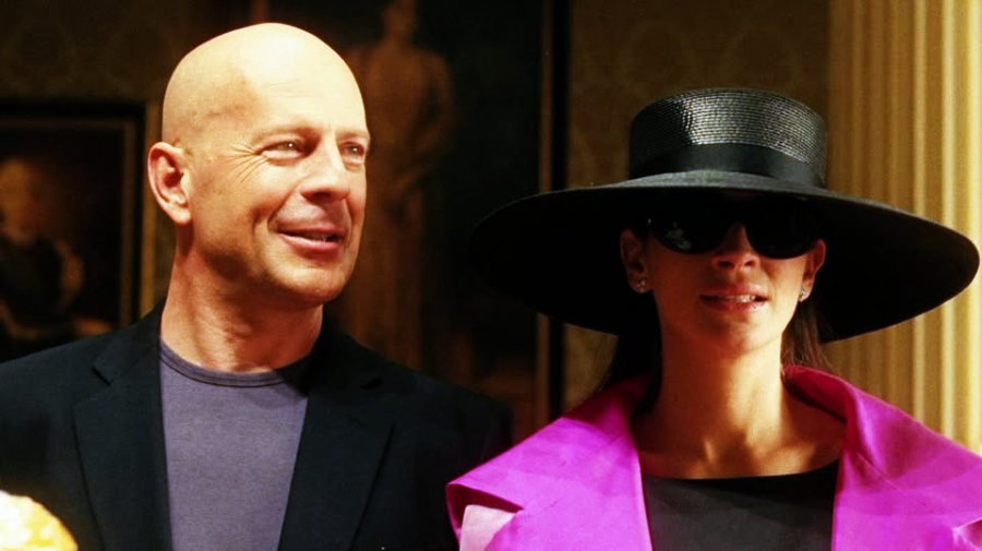 A bald man in a dark blazer stands smiling next to a woman wearing a large black hat, dark sunglasses, and a bright pink coat. They are indoors, with classical decor in the background.