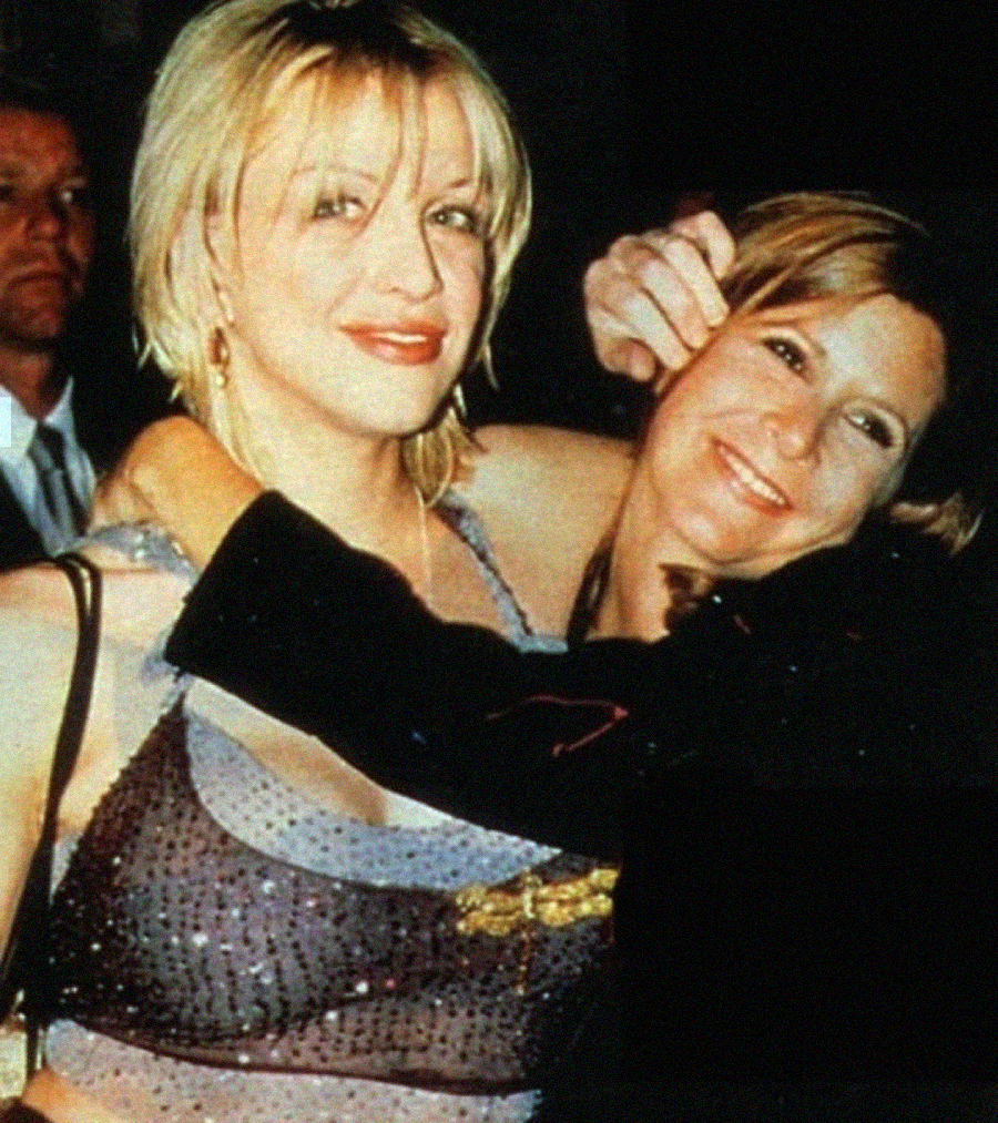 Two women pose together at an event, one with short blonde hair wearing a sheer top, and the other smiling with her arm around the first woman. A man in a suit stands blurred in the background.