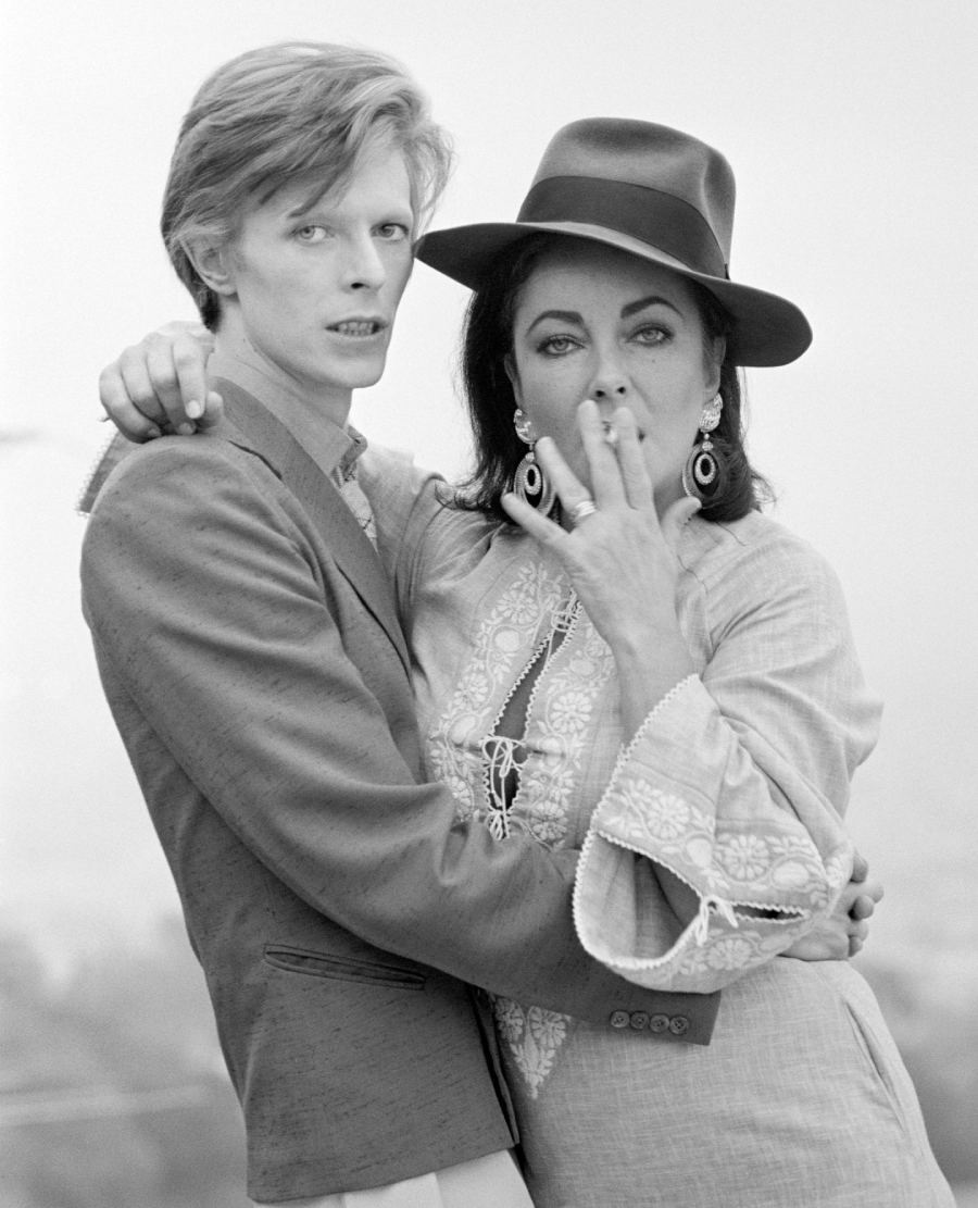 A black-and-white photo of a person with short hair in a blazer standing close to another person in a hat and embroidered dress, who is holding a hand to their mouth as if exhaling smoke.