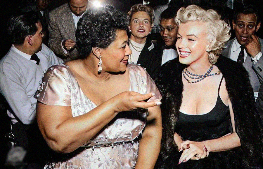 Two women, one in a light pink dress and the other in a black dress with a fur stole, smile and chat at a lively social event, surrounded by people in formal attire.