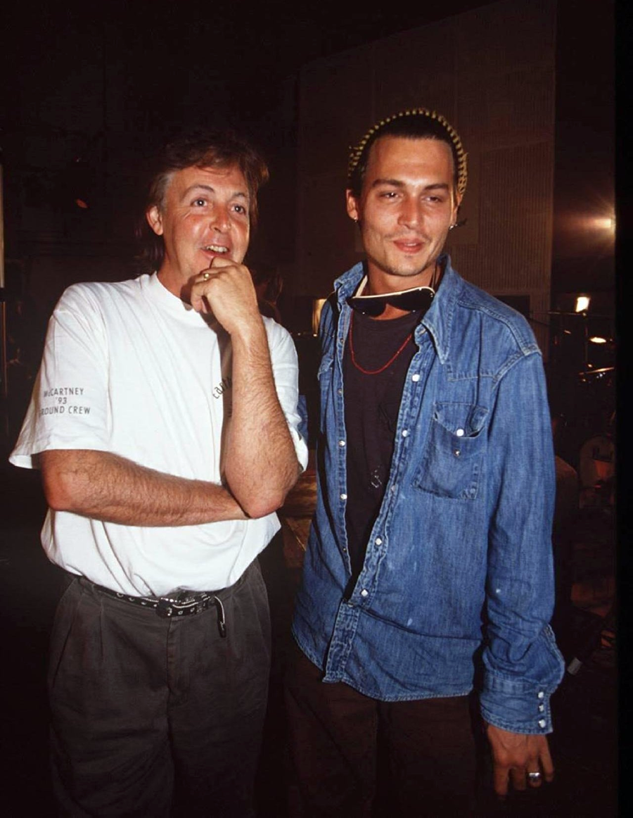 Two men stand together indoors; one wears a white t-shirt and dark pants, with his hand on his chin, while the other wears a denim shirt, a knit hat, and necklaces, smiling slightly. The background is dimly lit.