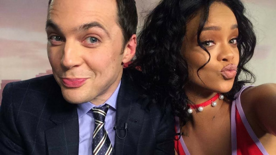 Two people take a close-up selfie. The person on the left is wearing a suit and tie, smiling with pursed lips. The person on the right has curly hair, is wearing a necklace, and is also making a pouty face.