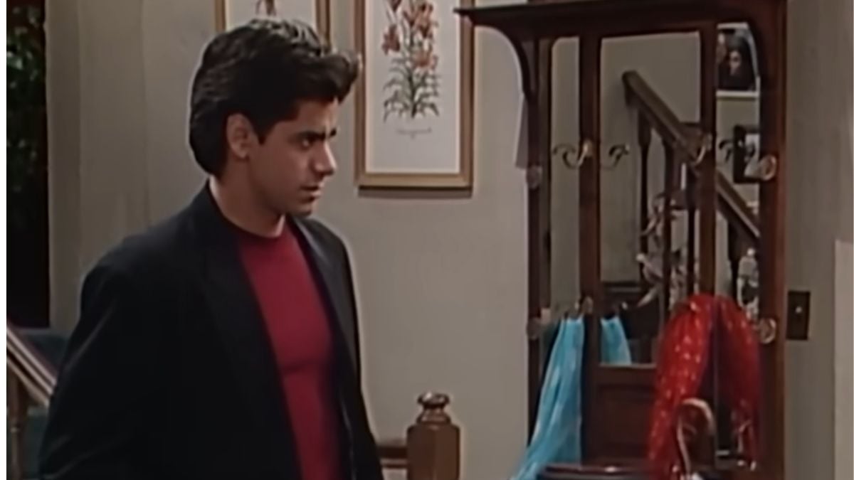 A man with dark hair in a red shirt and black blazer stands indoors near a coat rack with blue and red scarves. Framed floral pictures hang on the wall behind him.