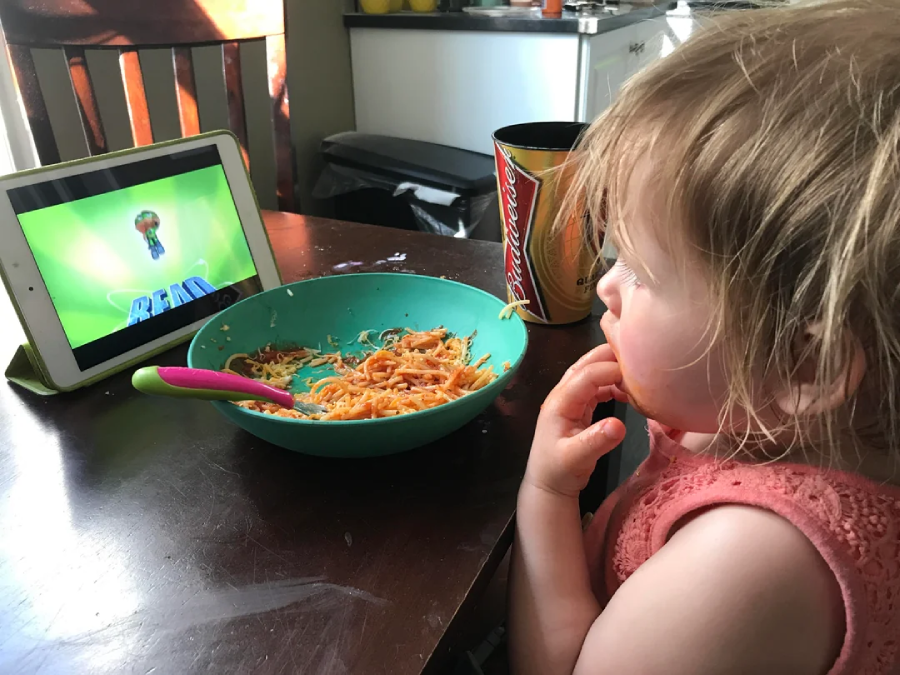 A young child with spaghetti sauce on their face watches a tablet while eating spaghetti from a bowl at a table. A colorful spoon rests in the bowl, and a cup is nearby.