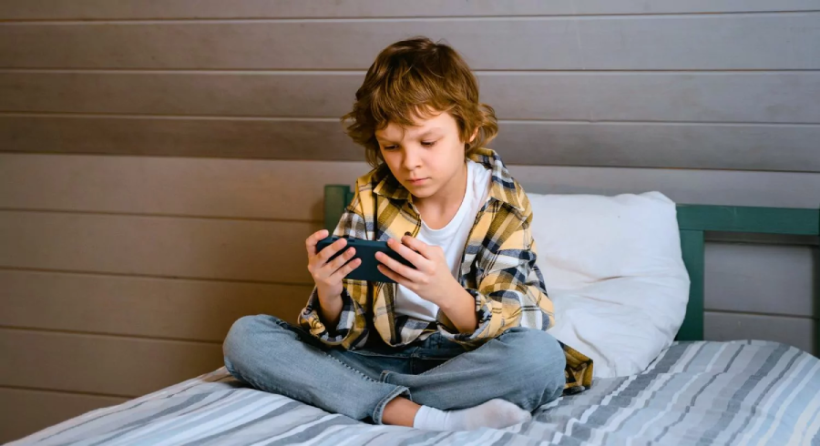 A young boy sits cross-legged on a bed, intently looking at a smartphone he holds in both hands. He wears a yellow plaid shirt, jeans, and socks. The background shows a gray wooden wall.
