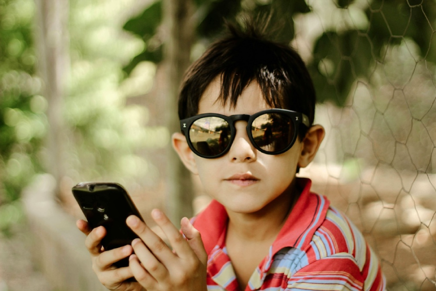 A young boy wearing dark sunglasses and a colorful striped shirt holds a smartphone while looking at the camera. He is outdoors, with greenery and a wire fence in the background.