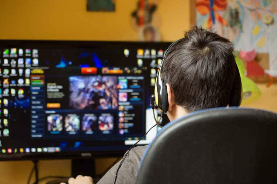 A person wearing headphones sits at a desk, facing a computer screen displaying a colorful video game interface, with icons and artwork visible. The room has a yellow wall and various decorations.