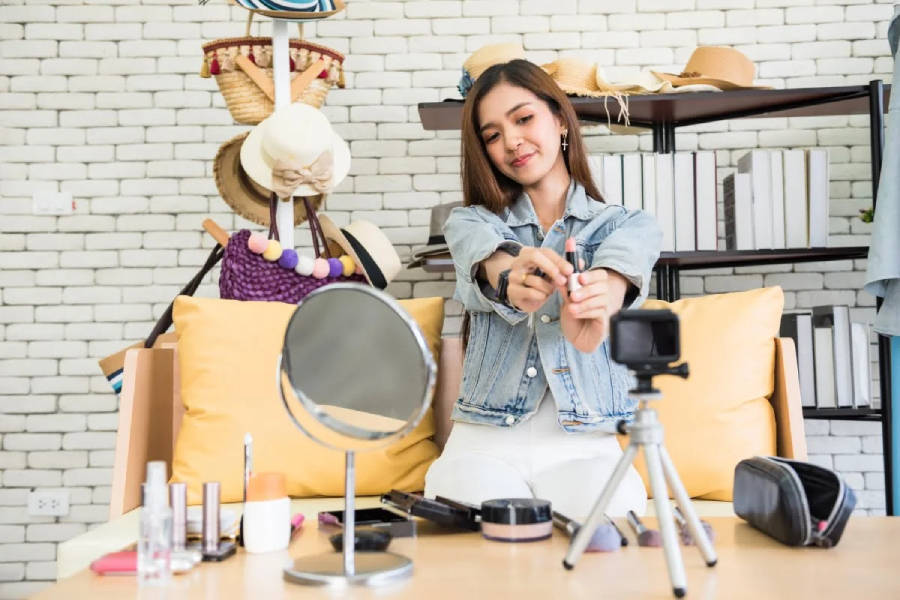 A young woman sits on a sofa, smiling while applying makeup in front of a camera and a mirror. Makeup products are spread on the table, and hats hang on the wall behind her.