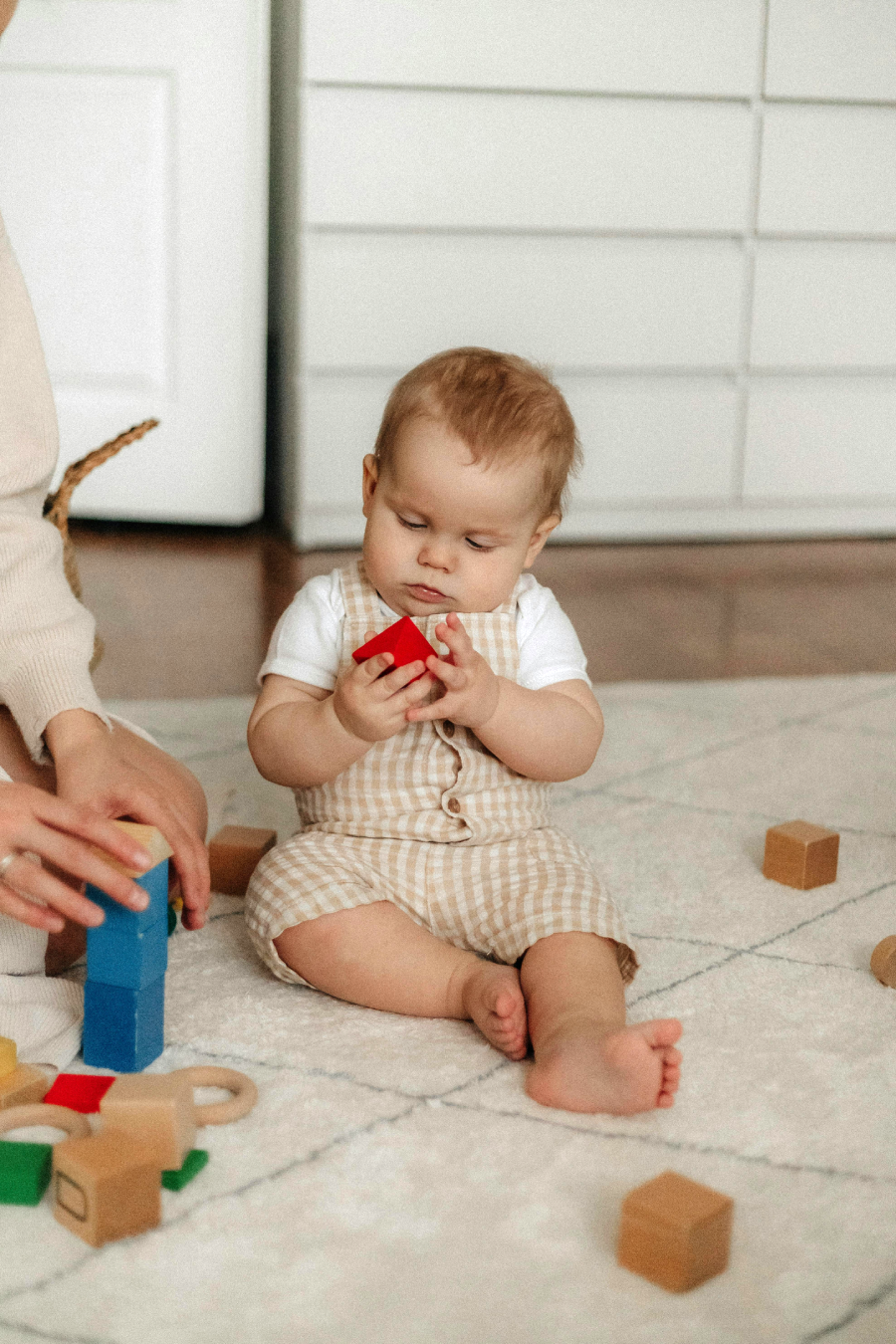 A baby in checkered overalls sits on a light carpet, holding a red toy block, surrounded by wooden blocks. An adult’s hands stack colorful blocks nearby. White drawers are in the background.