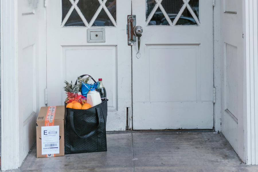 A black reusable bag filled with groceries and a cardboard package sits on a doorstep in front of white double doors with glass panes.