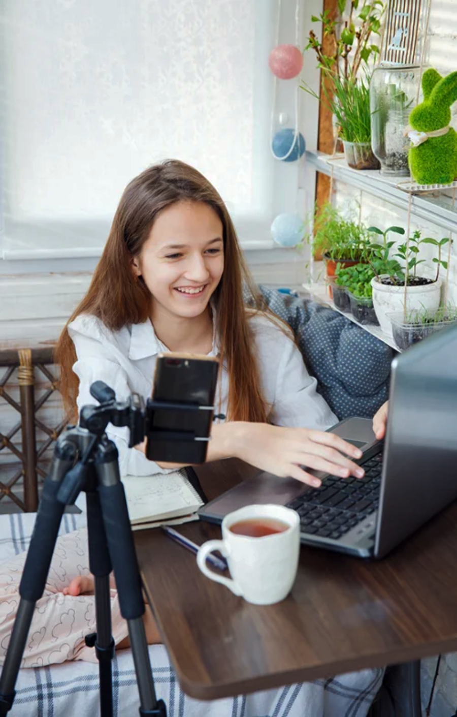 A smiling girl sits at a table using a laptop, with a smartphone mounted on a tripod in front of her. A cup of tea and a notebook are on the table, and plants are visible in the background.