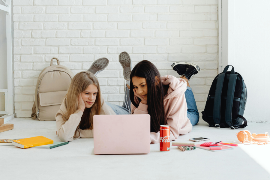 Two teenage girls lie on the floor in front of a pink laptop, surrounded by school supplies, notebooks, backpacks, and a soda can, working or studying together in a bright room with a white brick wall.