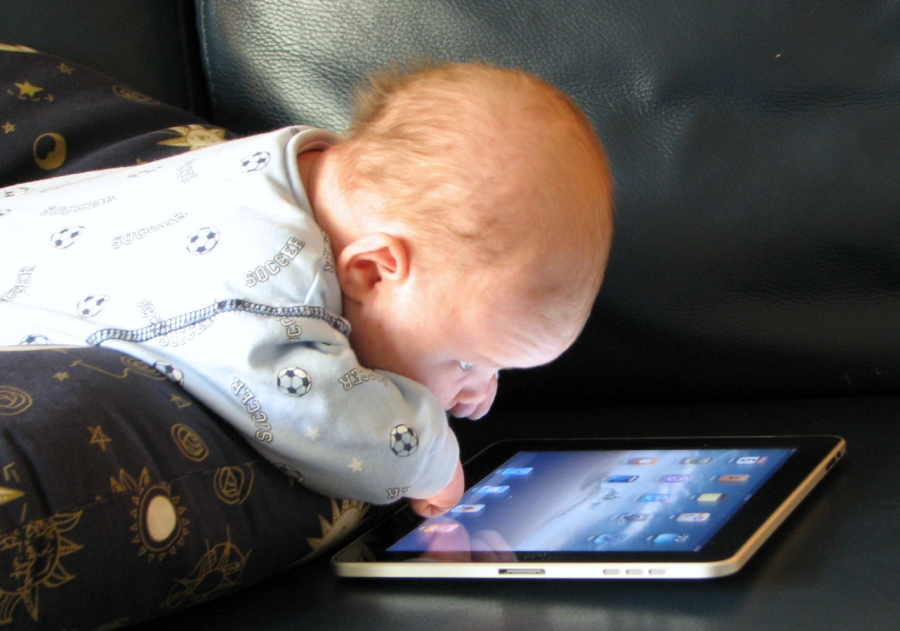 A baby wearing a blue onesie lies on a pillow, intently touching and looking at a tablet with its screen lit up, on a dark couch.