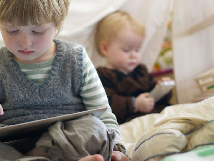 Two young children sitting indoors, each focused on a tablet device. The child in the foreground wears a striped shirt and gray vest, while the child in the background has blond hair and is blurred. Blankets and bedding surround them.