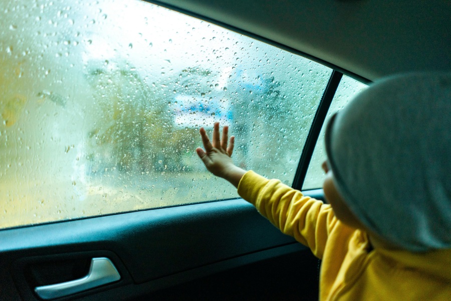 A child wearing a yellow sweater and gray beanie touches a rainy car window, looking outside at blurred scenery through water droplets on the glass.