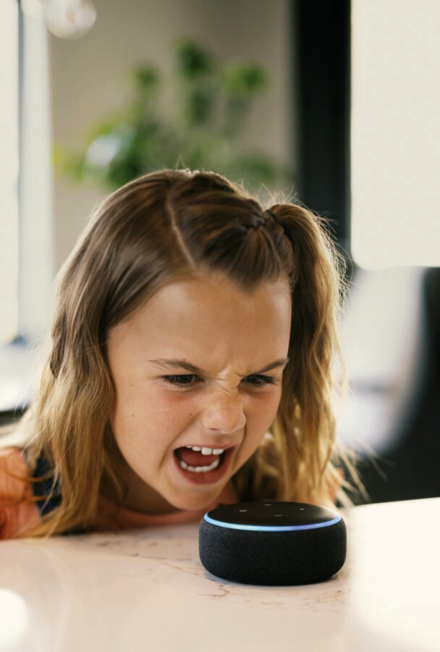 A young girl with long hair leans over a table, angrily shouting at a small black smart speaker with a blue ring, indoors with a blurred background.