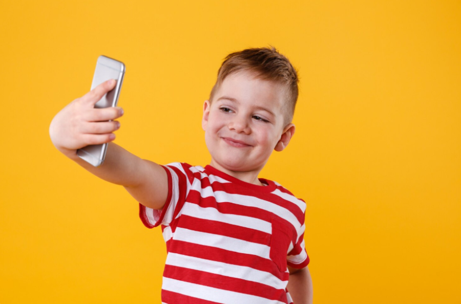 A young boy in a red and white striped shirt smiles while taking a selfie with a smartphone against a solid yellow background.